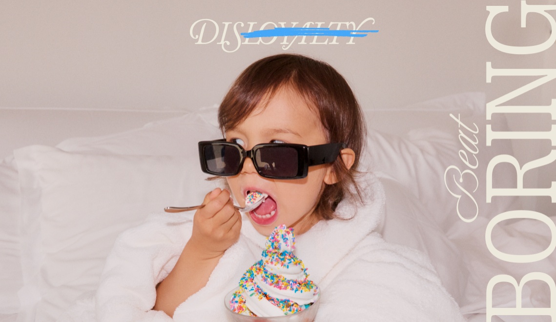 Et barn i en fluffy hvid badekåbe med solbriller nyder en skål med en dessert toppet med farverige krymmel, med en legende tekstoverlay.