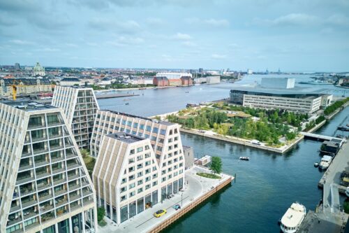 Luftfoto af moderne bygninger langs en vandvej med grønne områder, både og byens skyline i København under en overskyet himmel.
